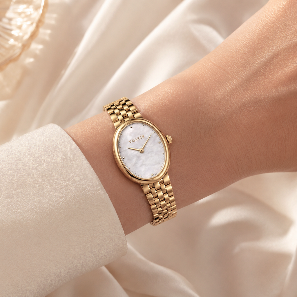 A gold-tone wristwatch with a white mother-of-pearl oval dial and matching gold link bracelet, shown worn on a woman's wrist against a soft cream satin background.
