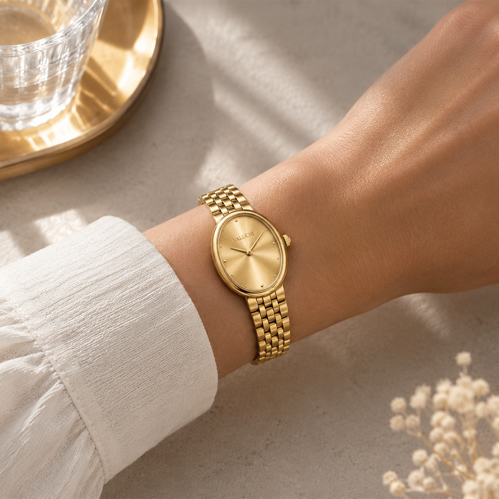 A gold-tone analog watch with a matching metal link bracelet worn on a person's wrist, shown against a neutral textured surface with soft shadows and a glass on a tray in the background.